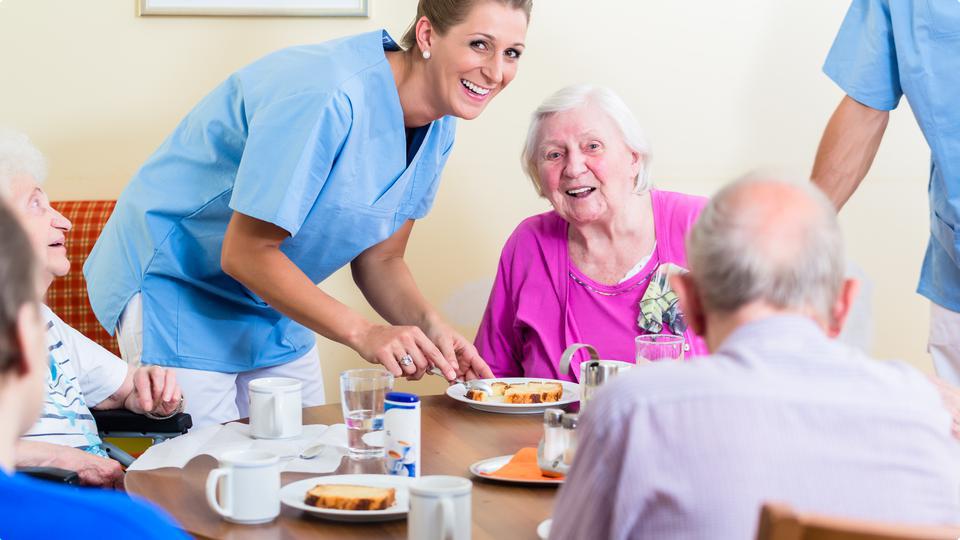 En grupp glada människor på ett äldreboende sitter vid ett runt bord och äter. En sköterska hjälper till.