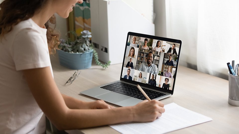 Kvinna deltar i ett digitalt möte. Sitter vid ett skrivbord med en bärbar dator framför sig. 